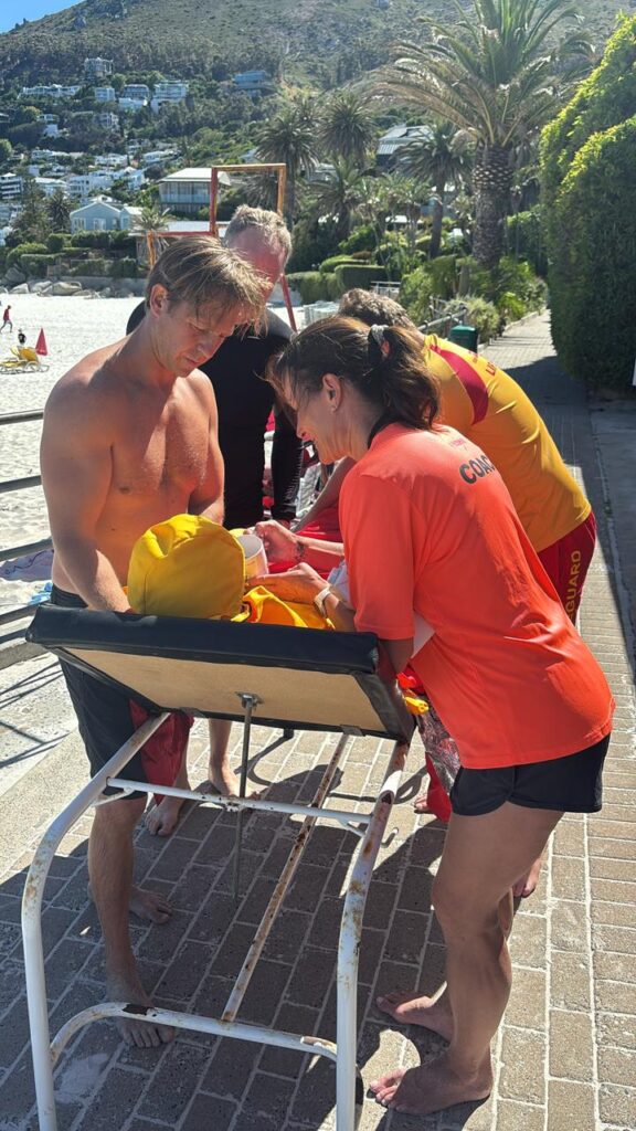 Clifton Lifeguards, Glenn, Tracey and Teagan help the patient keep warm in front of the Clifton 4th Beach boat shed.